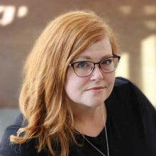 Image shows a smiling woman with long red hair and glasses wearing a black sweater